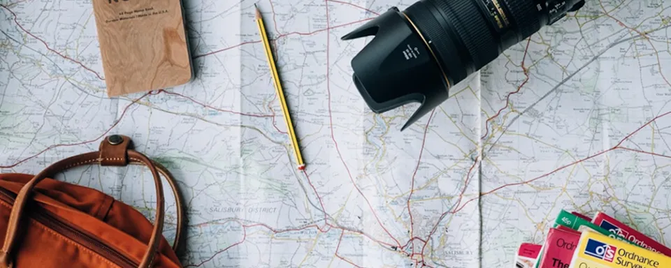 Folded map with a camera lens, pencil, notebook, and orange backpack arranged on top for travel planning.