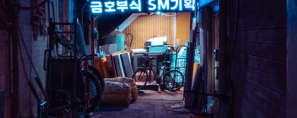 Dimly lit alleyway cluttered with bicycles, packages, and various stored items under a neon sign with Korean characters.