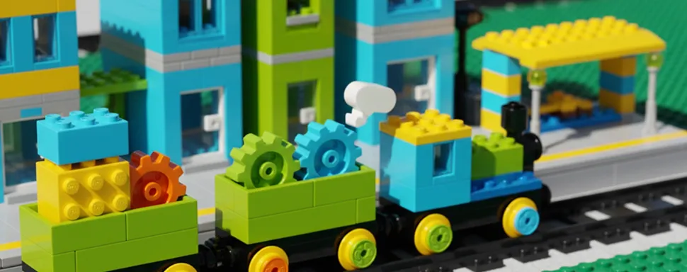 Toy train built from colorful interlocking plastic bricks, with blue, yellow, and green cars carrying gear-shaped bricks on black train tracks, set against a backdrop of blue and green brick buildings and a yellow and blue shelter.