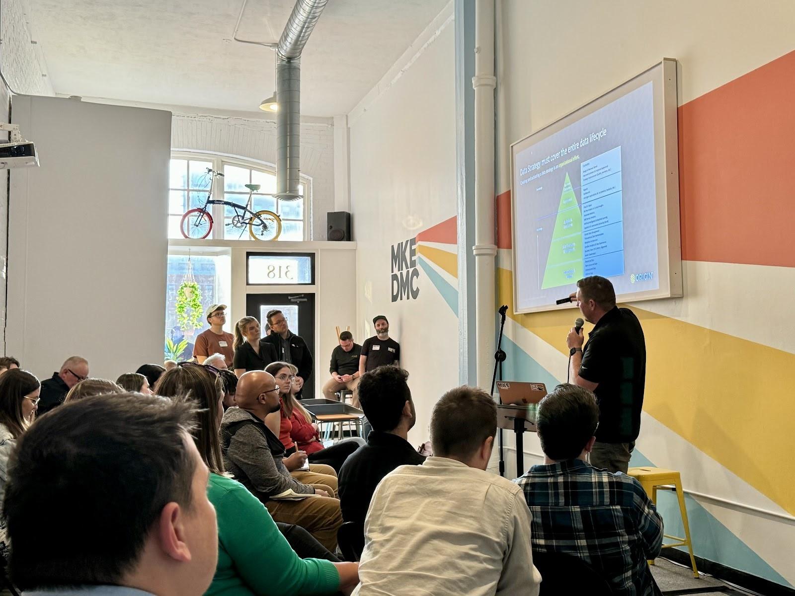 Nick White speaking in front of a screen with an infographic in front of a seated audience inside the MKE DMC clubhouse