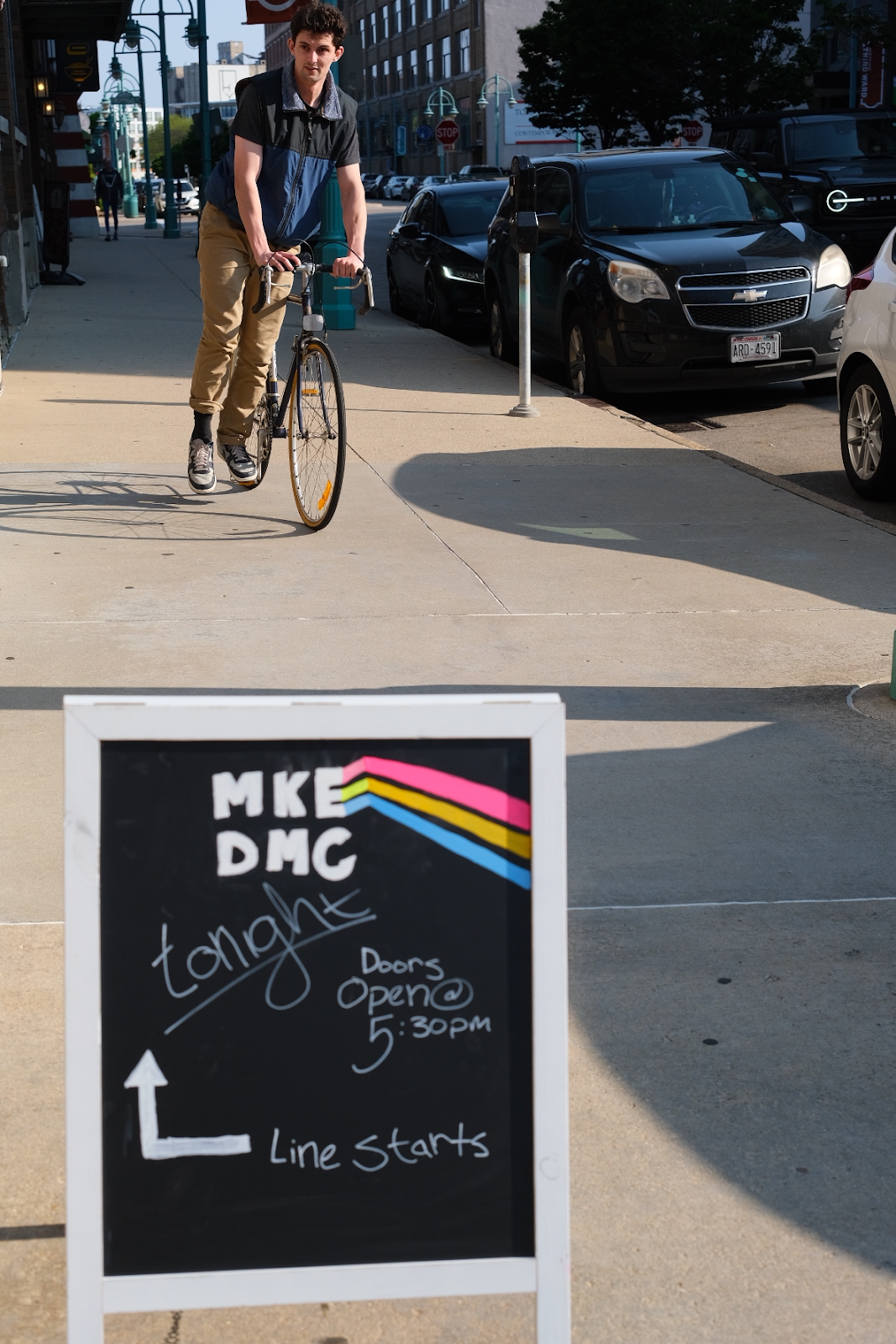  A man dismounting from a bicycle and a sandwich board announcing tonight’s MKE DMC event on the sidewalk in Milwaukee’s Historic Third Ward