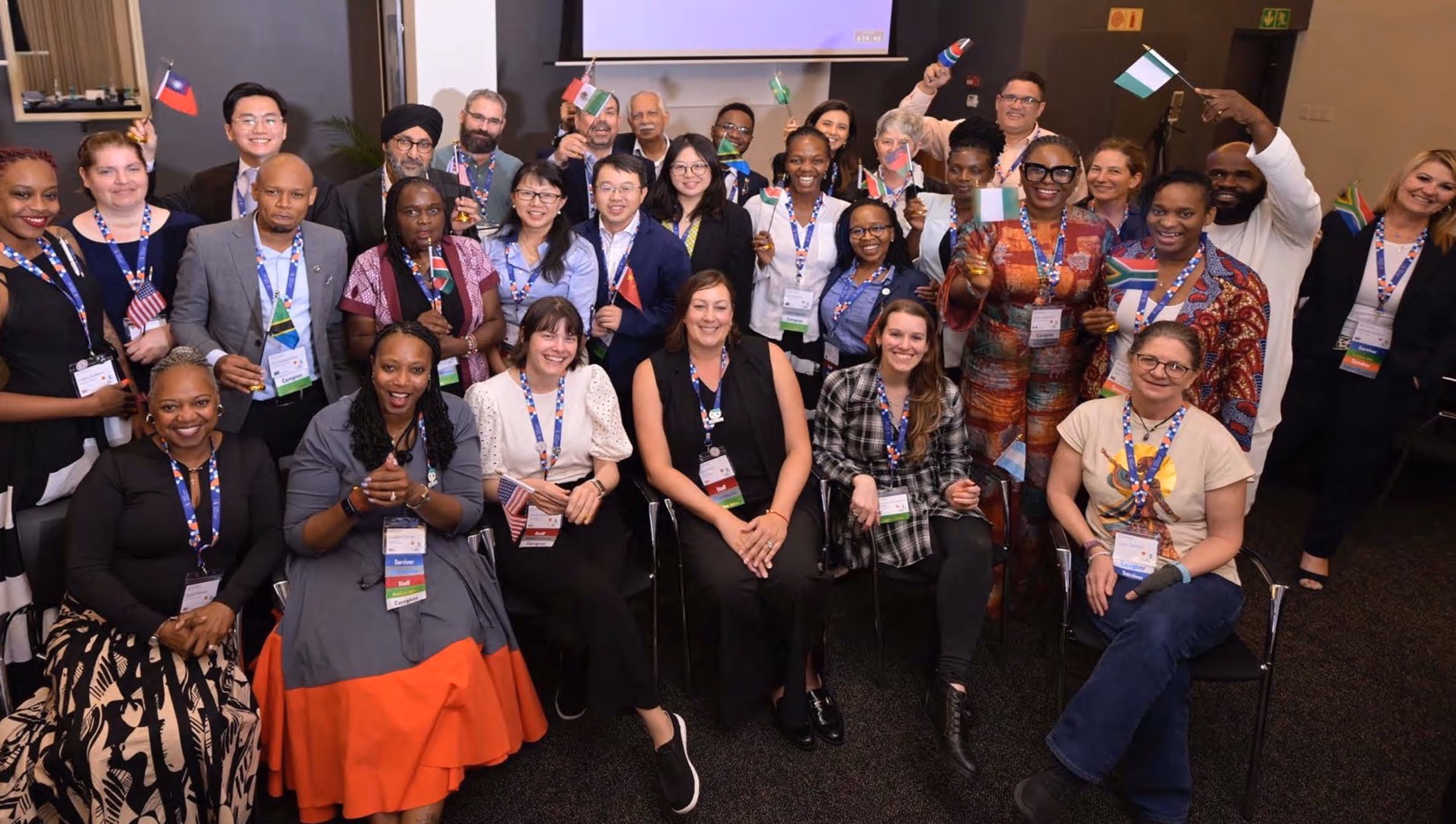 Group of diverse people smiling and seated and standing indoors holding various small international flags.