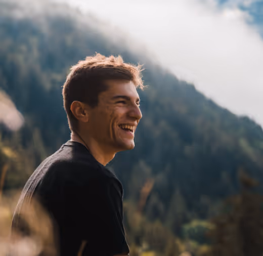 Young man with short brown hair wearing a black shirt smiling outdoors with forested mountains in the background.