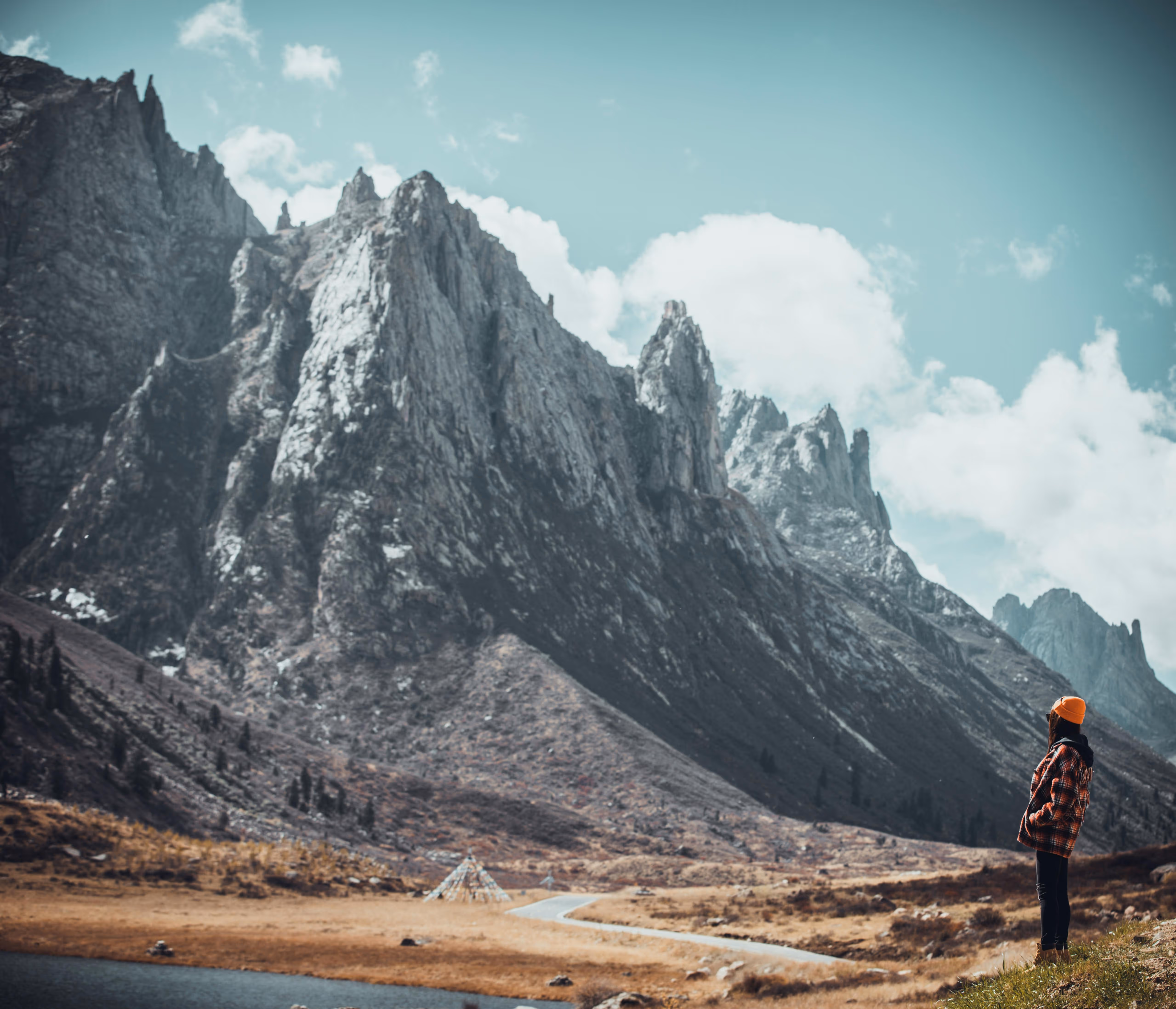Person wearing an orange beanie and plaid jacket standing on a hill overlooking a winding road and rocky mountain landscape under a blue sky with clouds.