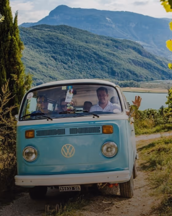 Group of four people inside a blue vintage Volkswagen van driving on a dirt road surrounded by greenery and mountains in the background.