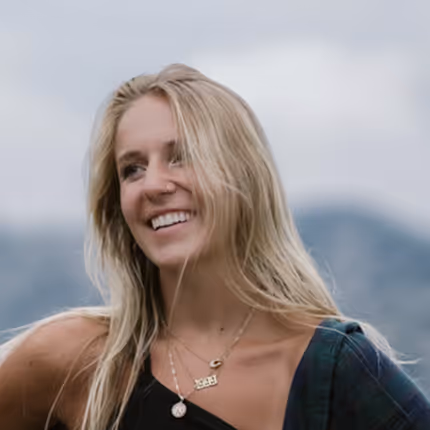 Smiling young woman with long blonde hair wearing necklaces and a one-shoulder top with a plaid shirt draped over one shoulder, outdoors with a blurred natural background.