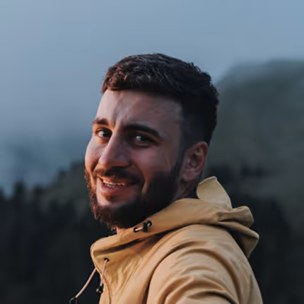Smiling man with short dark hair and beard wearing a mustard jacket outdoors with blurred forest background.