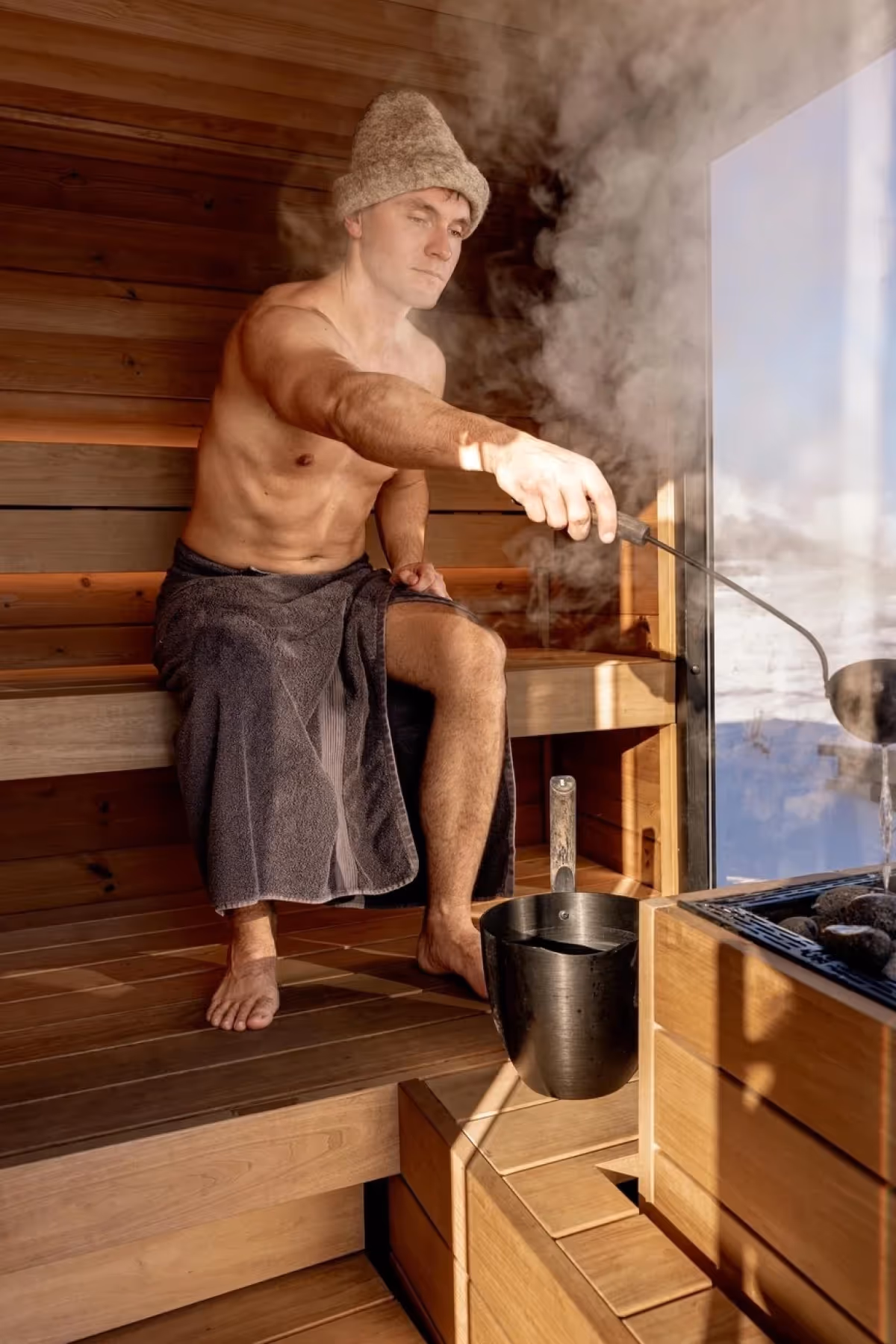 Man wearing a towel and hat sitting in a wooden sauna pouring water onto hot stones, creating steam.