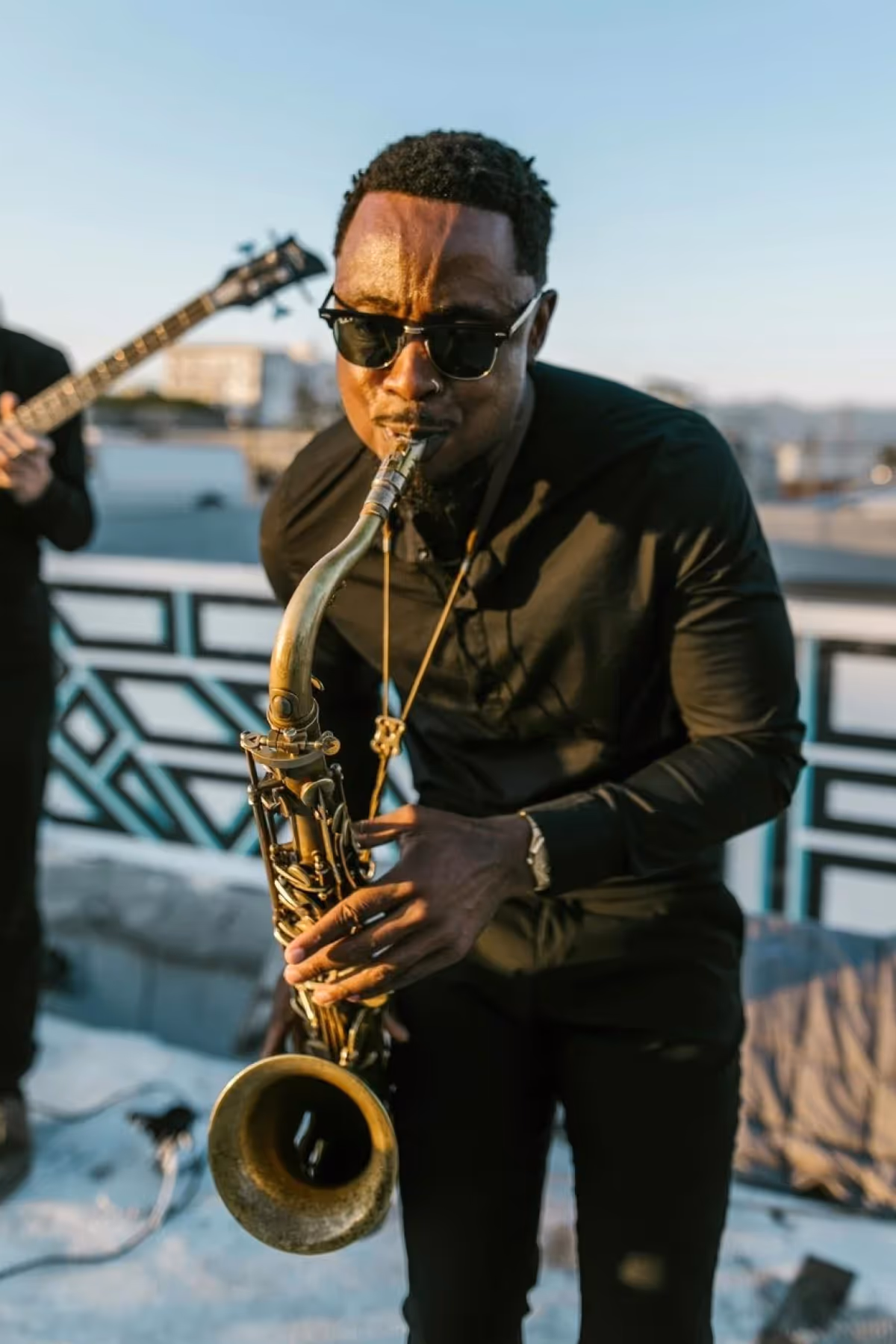 Man wearing sunglasses playing a saxophone outdoors in the sunlight.