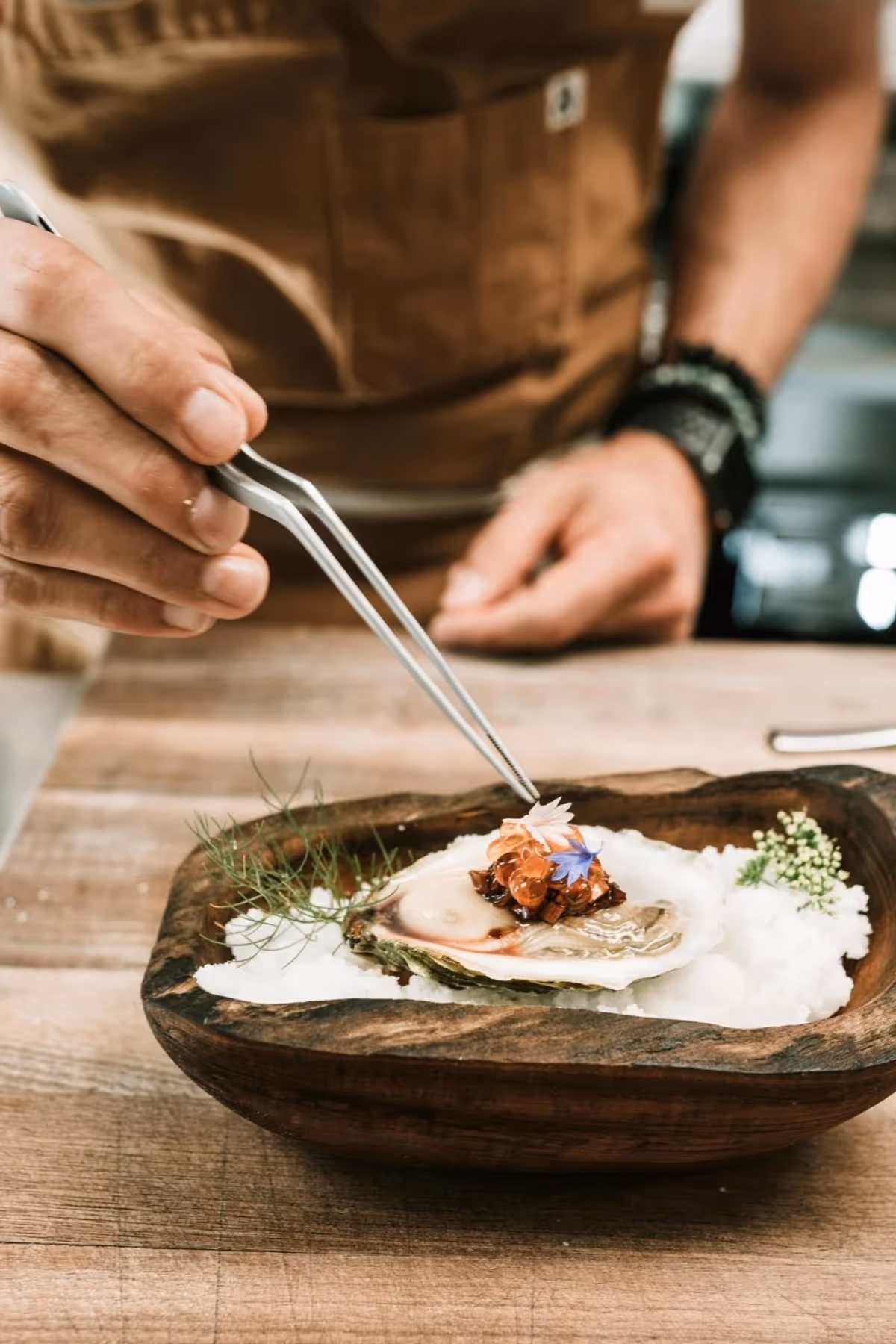 Chef using tweezers to garnish a decorated oyster on a wooden dish with ice and herbs.