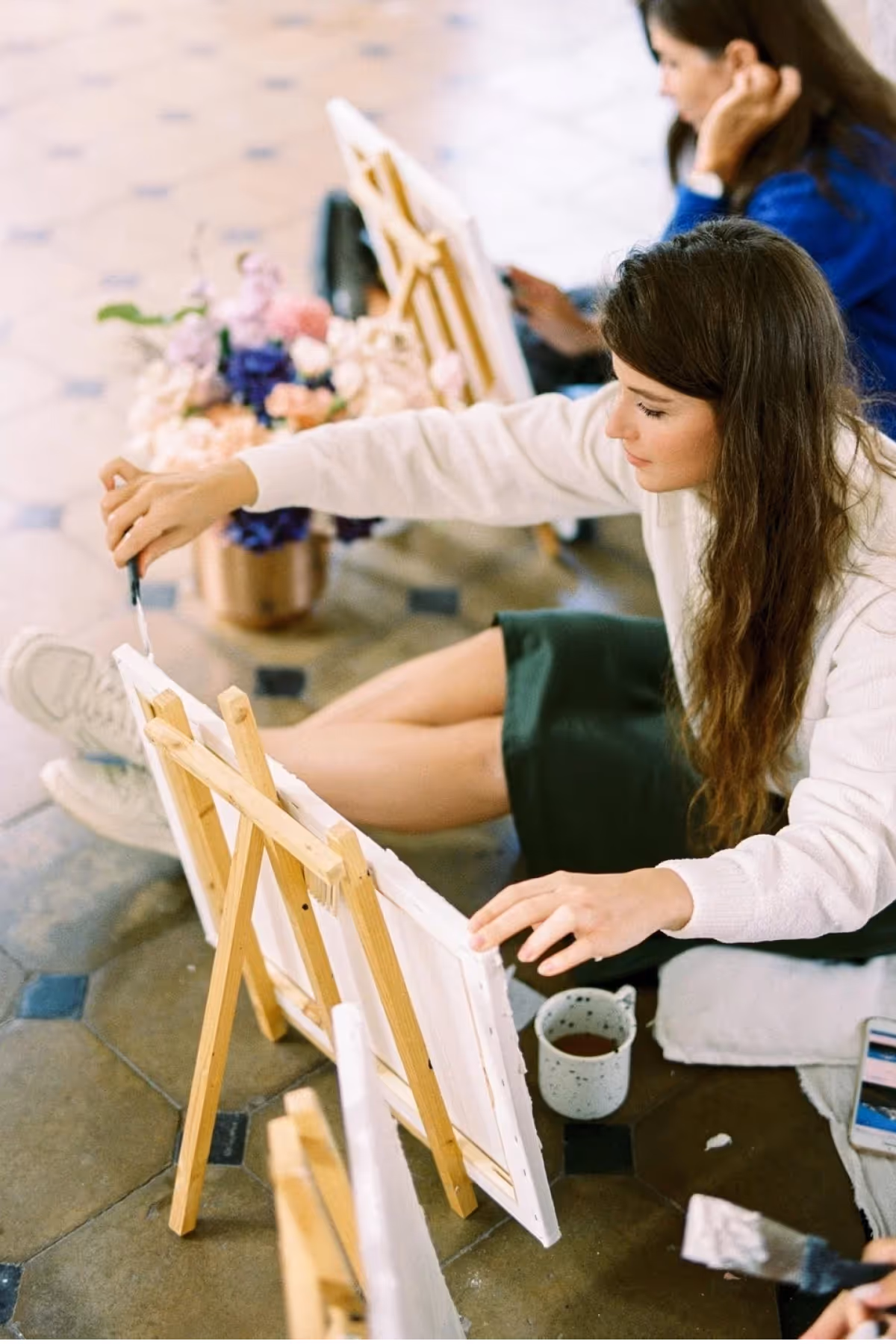 Young woman with long brown hair sitting on the floor painting on a canvas propped on a wooden easel, with a mug and flowers nearby.