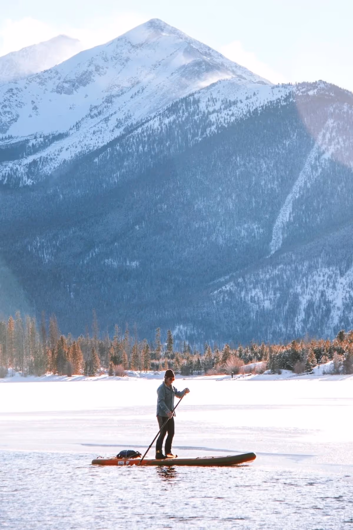 Person paddleboarding on a partially frozen lake with snow-covered mountains and pine trees in the background.