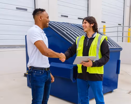 Business owner and waste service rep shaking hands near a commercial trash dumpster.
