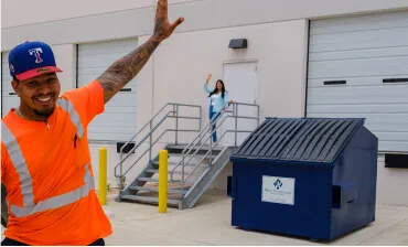 Waste service worker waving near a commercial trash dumpster at a business location.