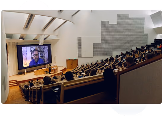Audience seated in a large lecture hall watching a speaker on a large screen at the front.