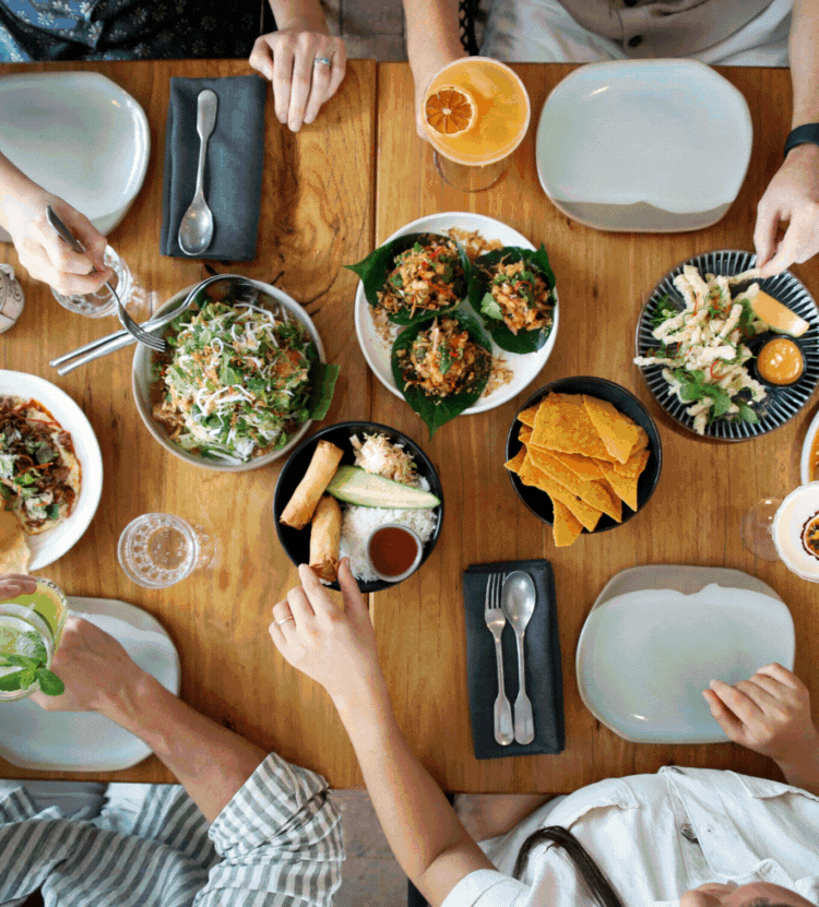Overhead view of a wooden table with various dishes including spring rolls, salad, chips, and drinks with four people seated around it.