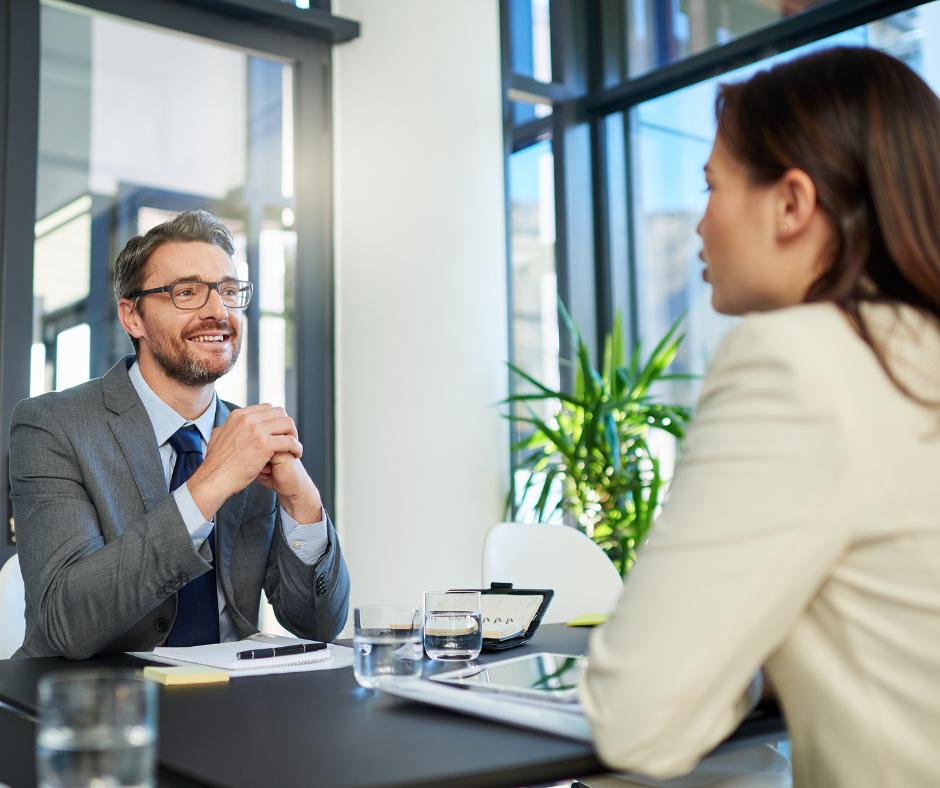 This image shows a professional man and woman sitting across from each other at a desk. The man is facing the viewer and smiling.