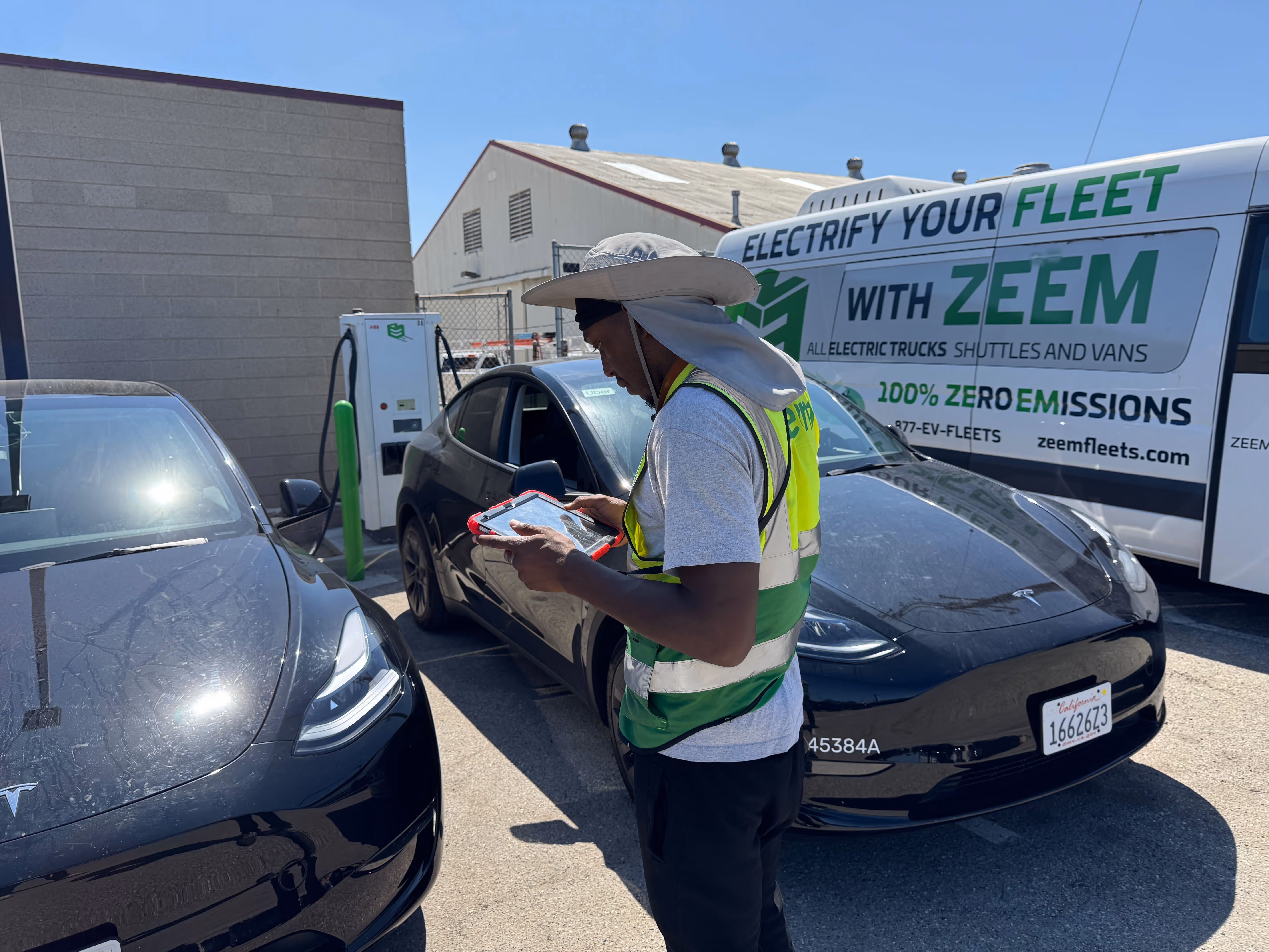 Worker wearing a high-visibility vest and wide-brimmed hat checking a tablet between two black Tesla electric cars near a ZEEM electric vehicle fleet van and charging station.