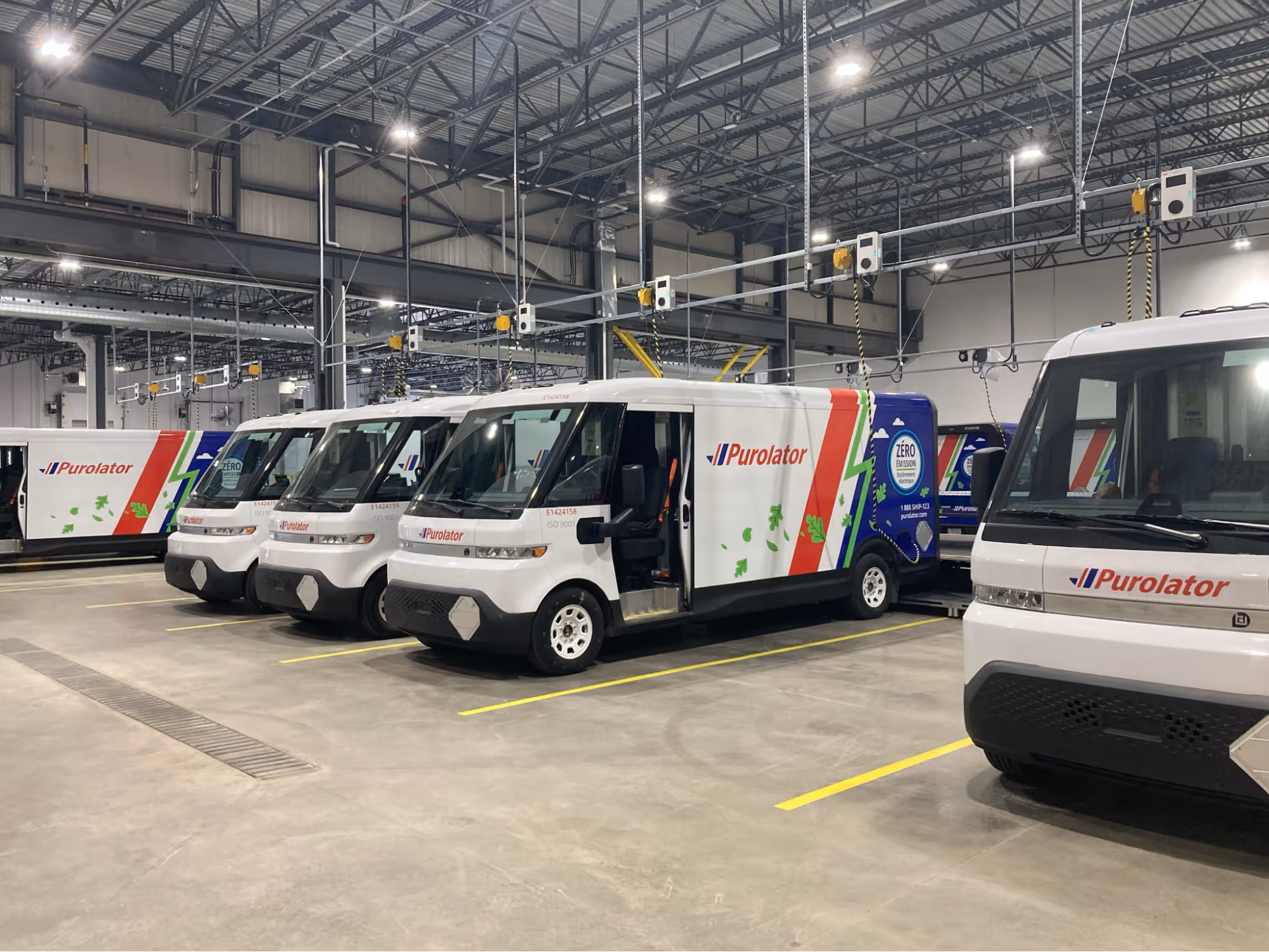 Indoor view of several white Purolator delivery electric vans parked in a warehouse with overhead charging stations.