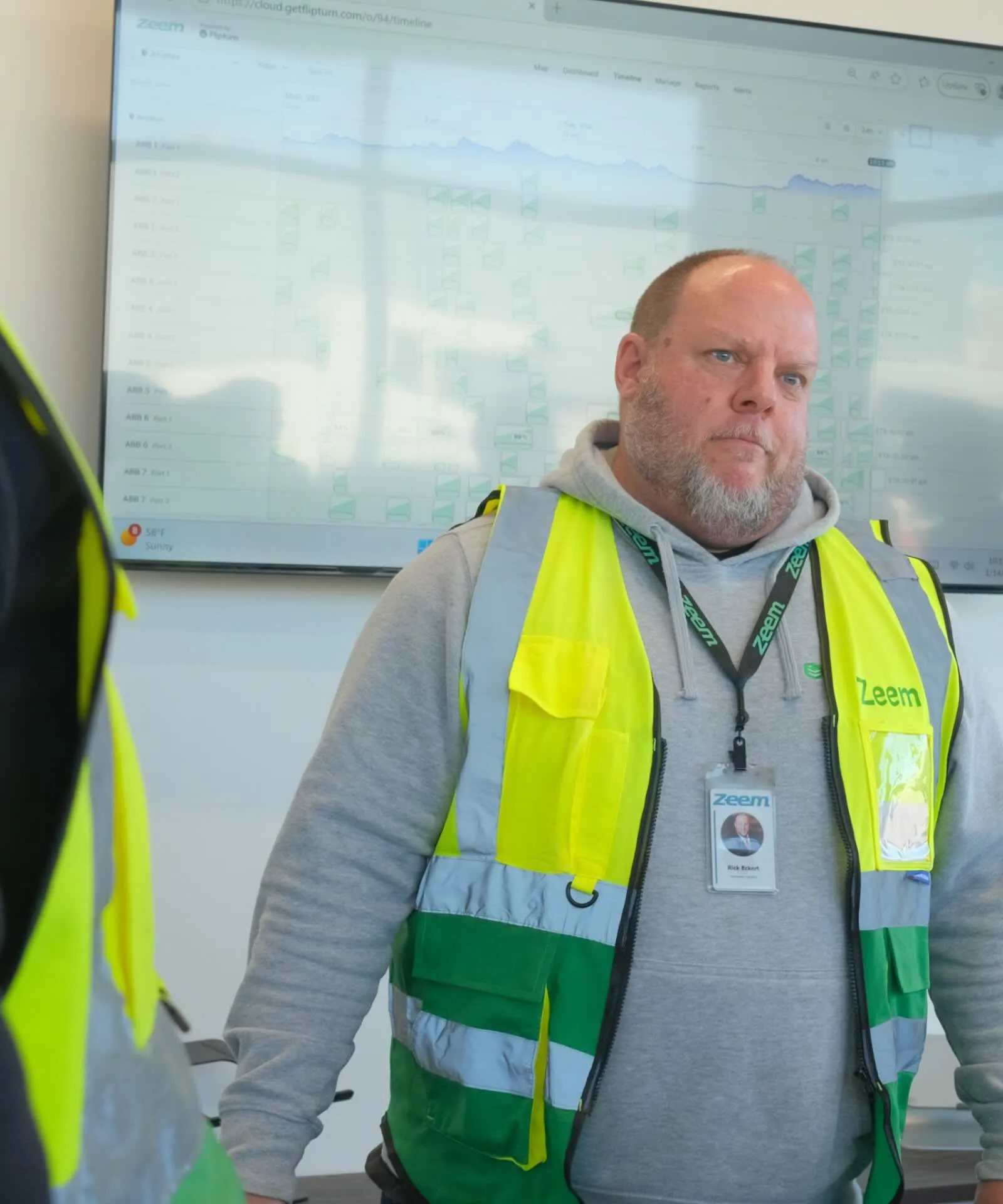 Man wearing a gray hoodie and a high-visibility yellow and green Zeem vest, standing indoors with a digital screen displaying charts in the background.