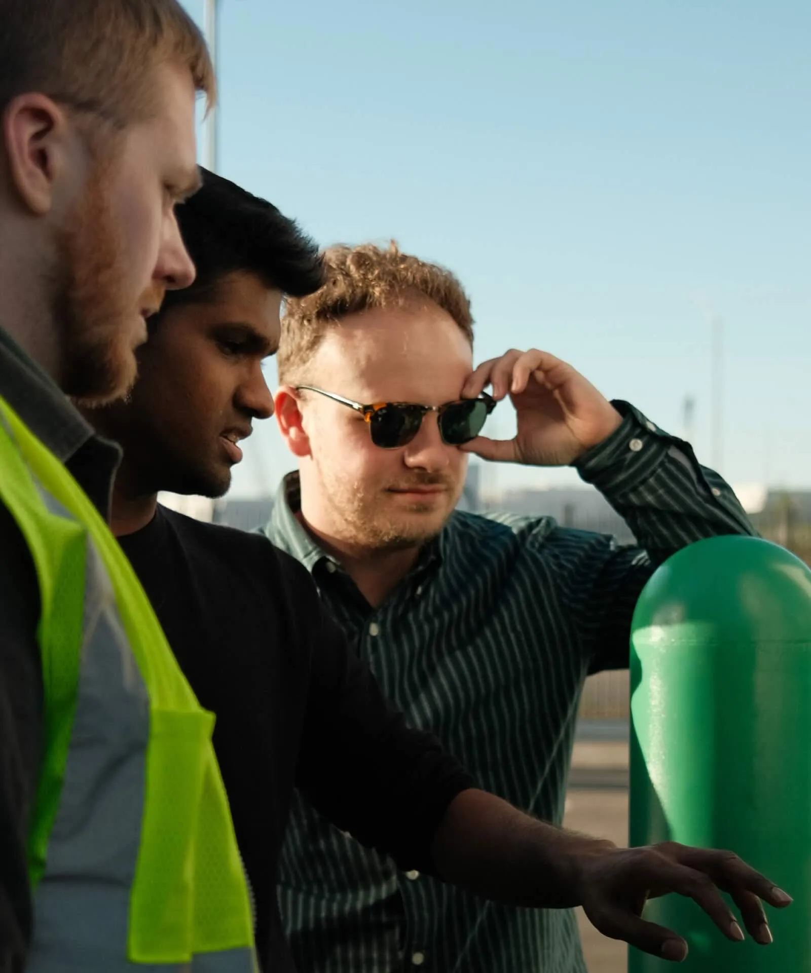 Three men closely examining something with one wearing a neon safety vest and another adjusting his sunglasses outdoors.