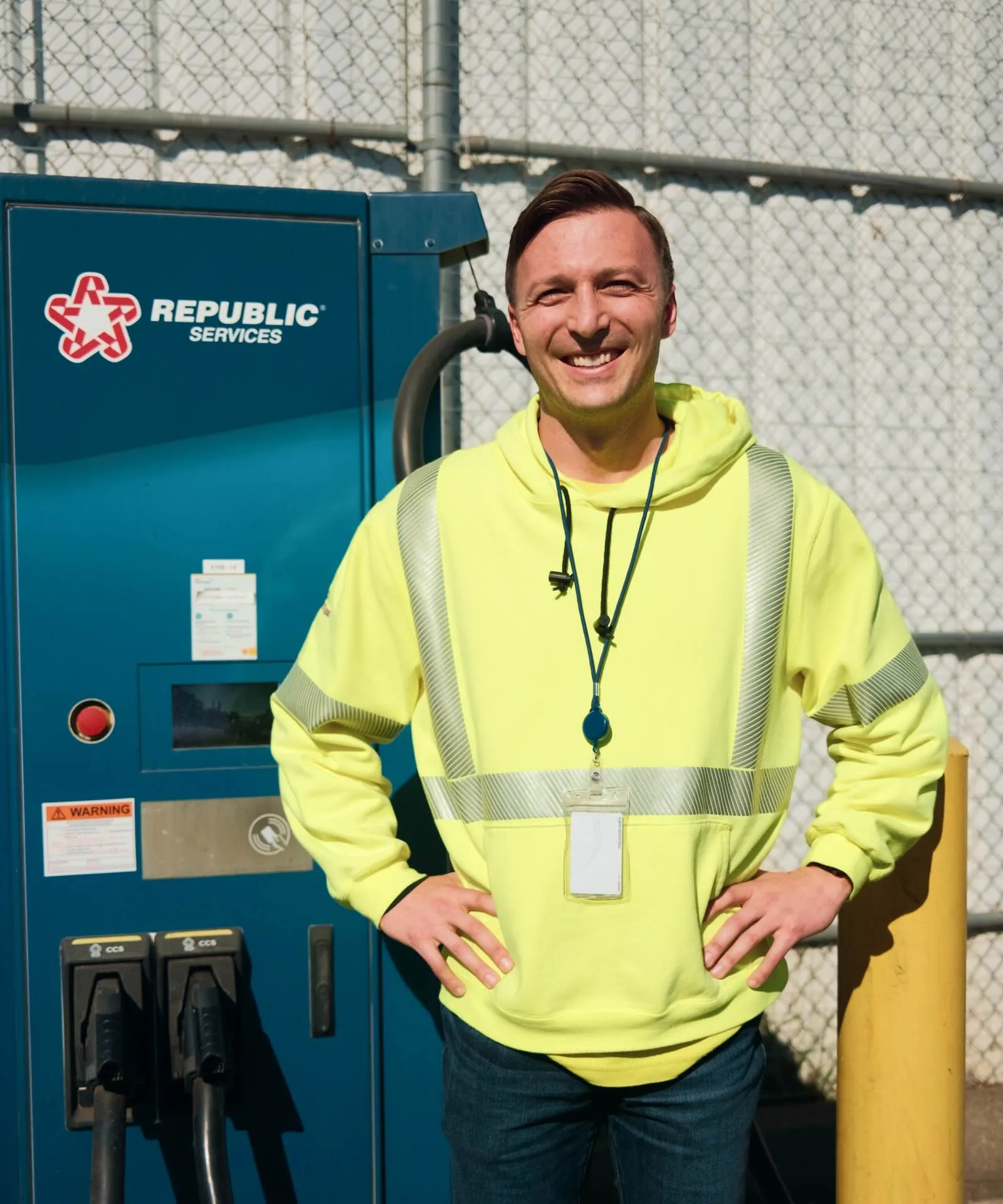 Smiling man in a yellow safety hoodie with reflective stripes stands with hands on hips in front of a Republic Services machine and chain-link fence.