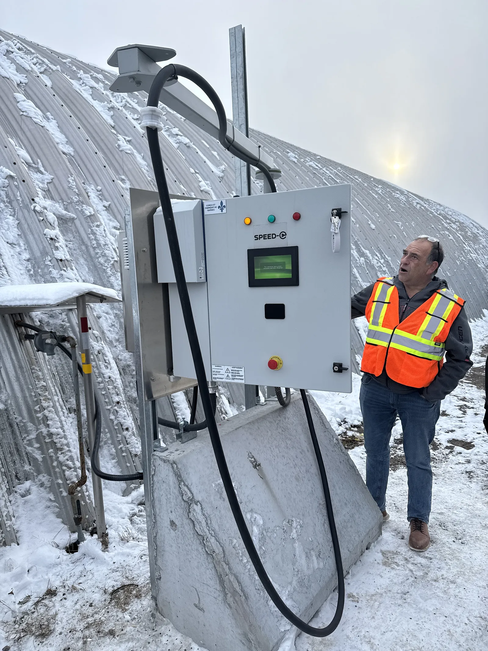 Man in an orange safety vest standing next to a SPEED-E electric control panel outdoors in a snowy environment.