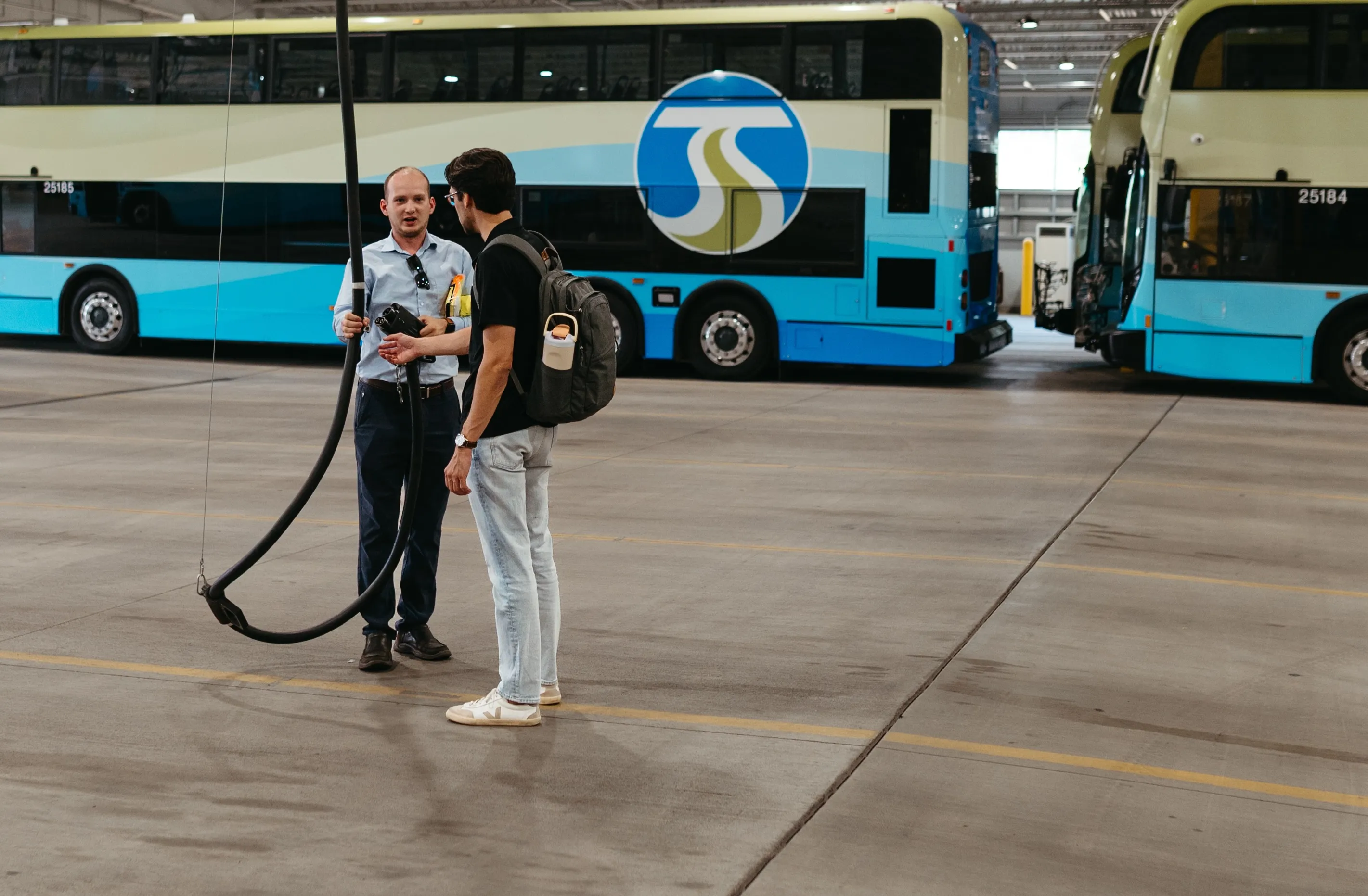 Two men talking inside a bus depot with blue and cream double-decker buses in the background.