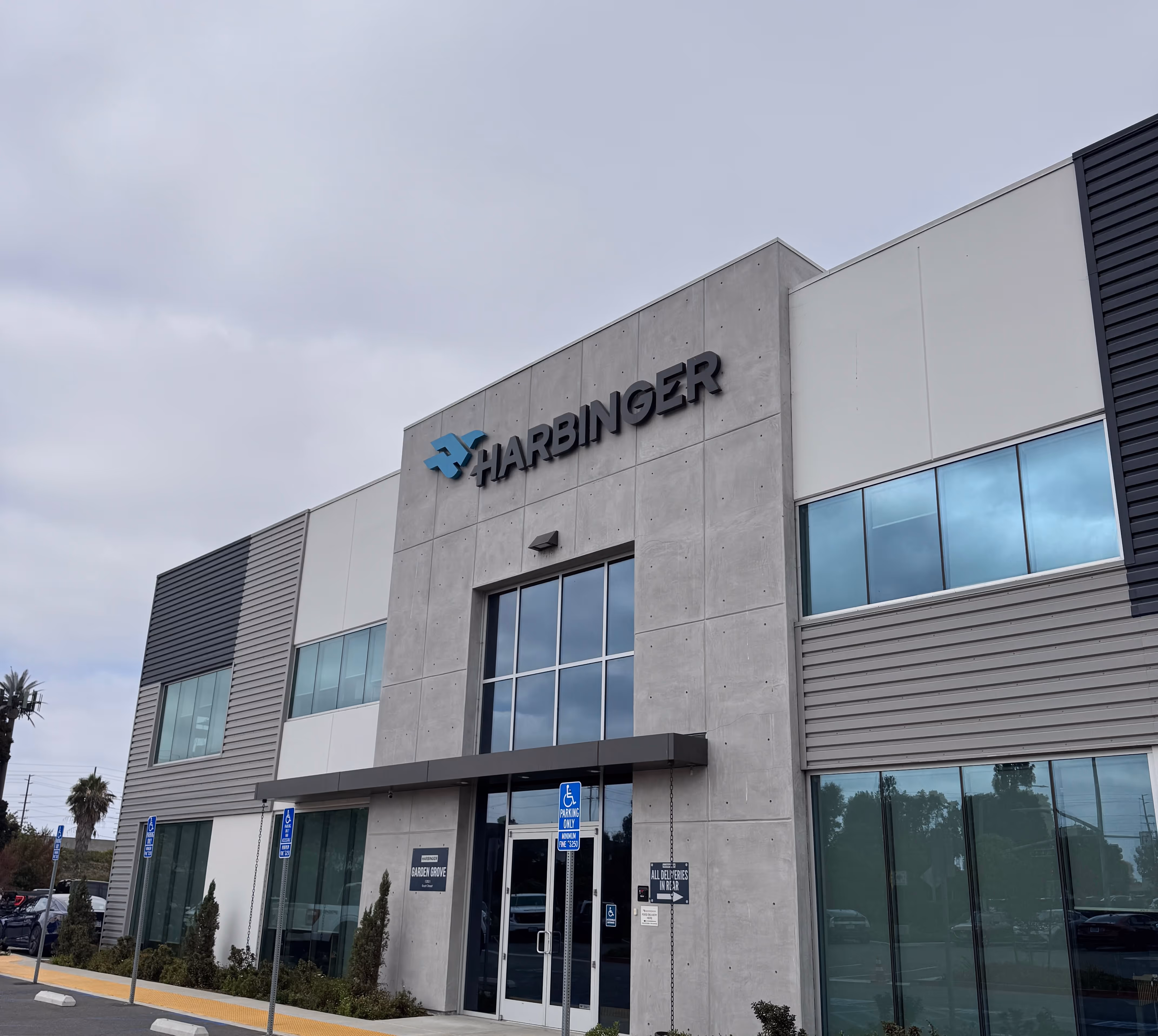 Modern two-story building with large windows and a sign reading 'Harbinger' above the entrance on a cloudy day.