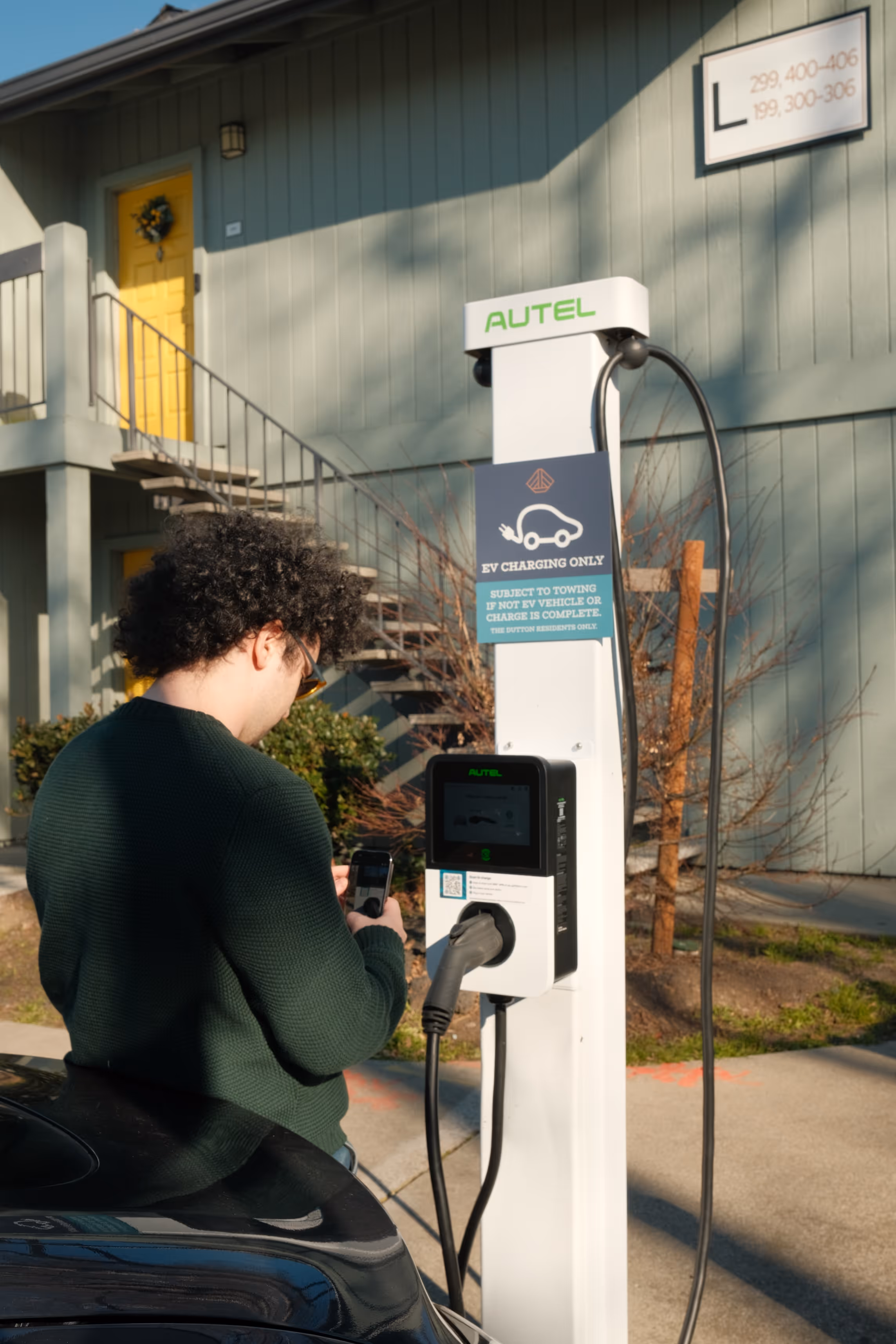 Person with curly hair using a smartphone next to an electric vehicle charging station in front of an apartment building with a yellow door.