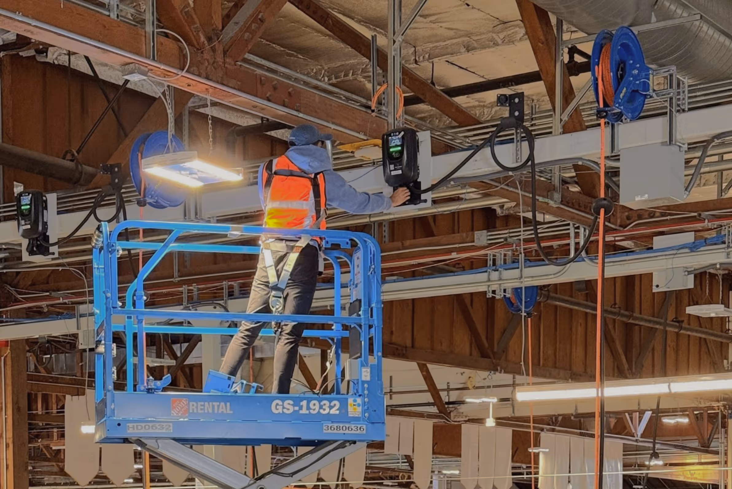 Worker in orange vest using a blue scissor lift to reach ceiling EV charging equipment in a large warehouse.