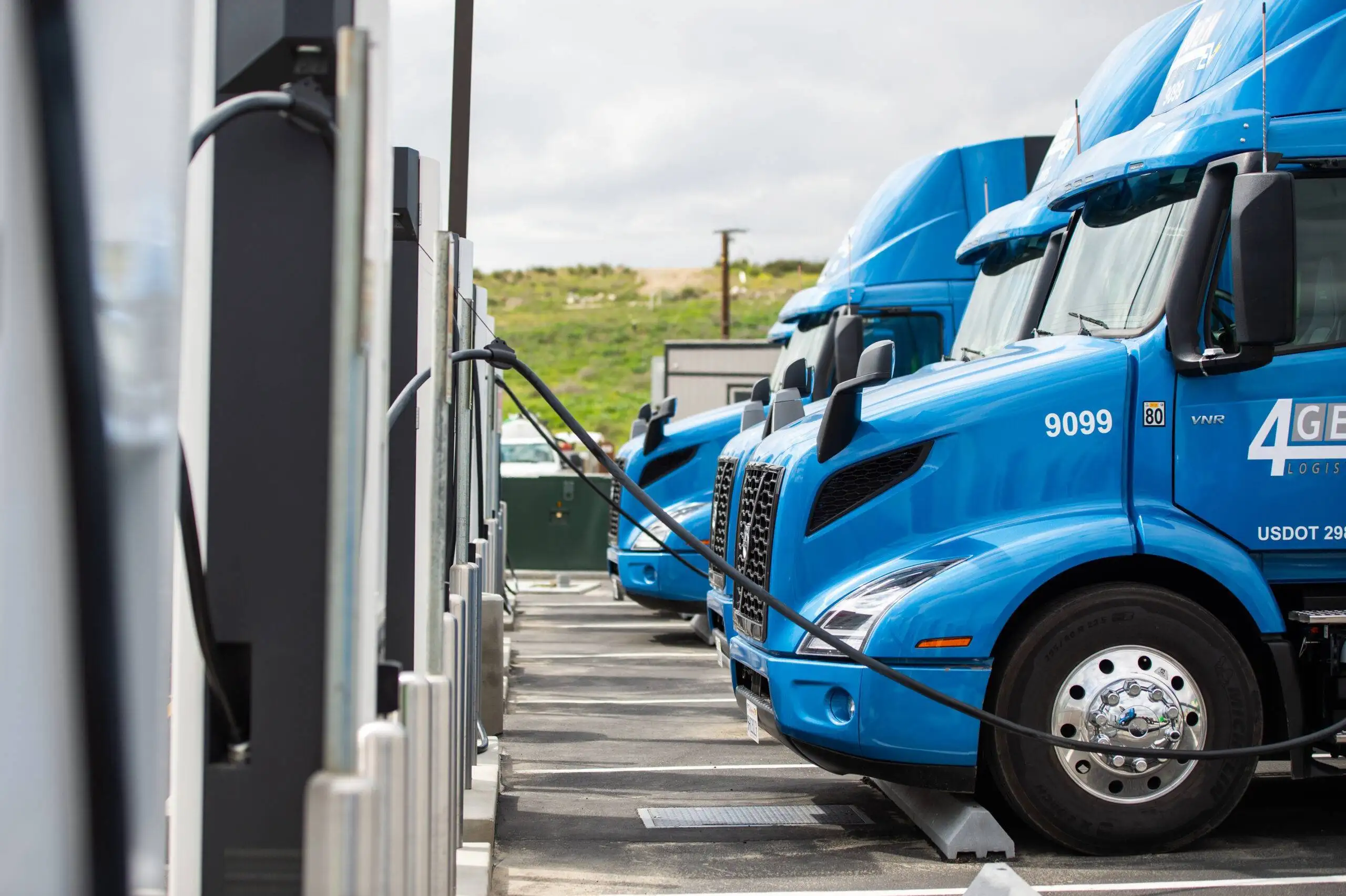 Row of blue electric trucks plugged into charging stations in an outdoor lot.