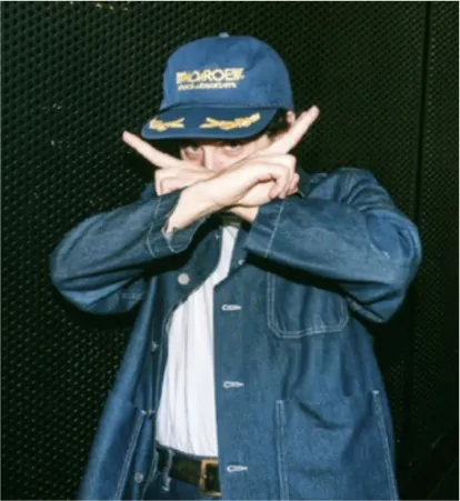 Person in a blue cap and denim jacket making an 'X' gesture with their arms in front of their face, partially obscuring their eyes. The cap reads 'MONROE AUTO EQUIPMENT' with yellow embroidery.