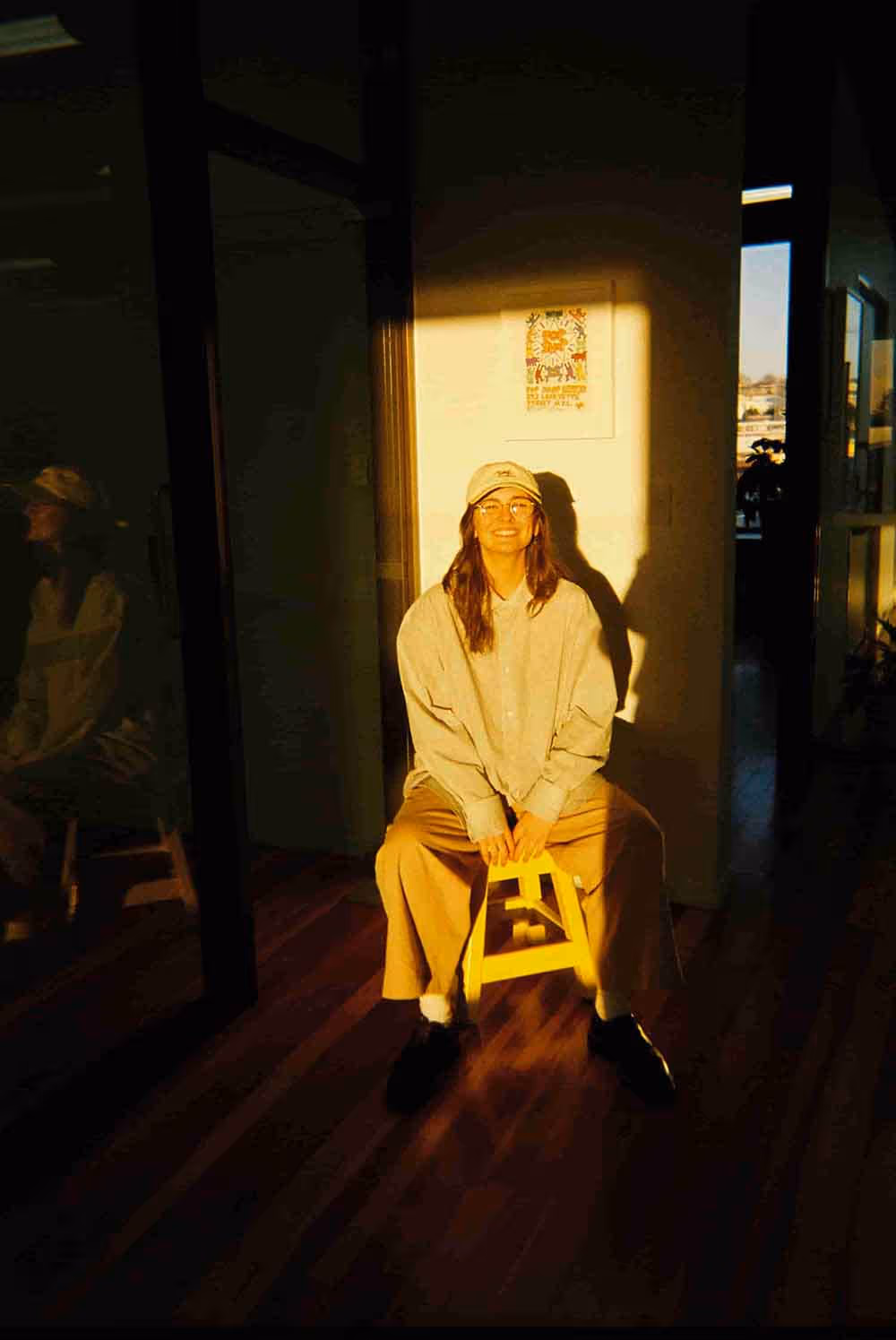 A person sitting on a stool in golden-hour light, smiling in the hallway of a creative workspace.