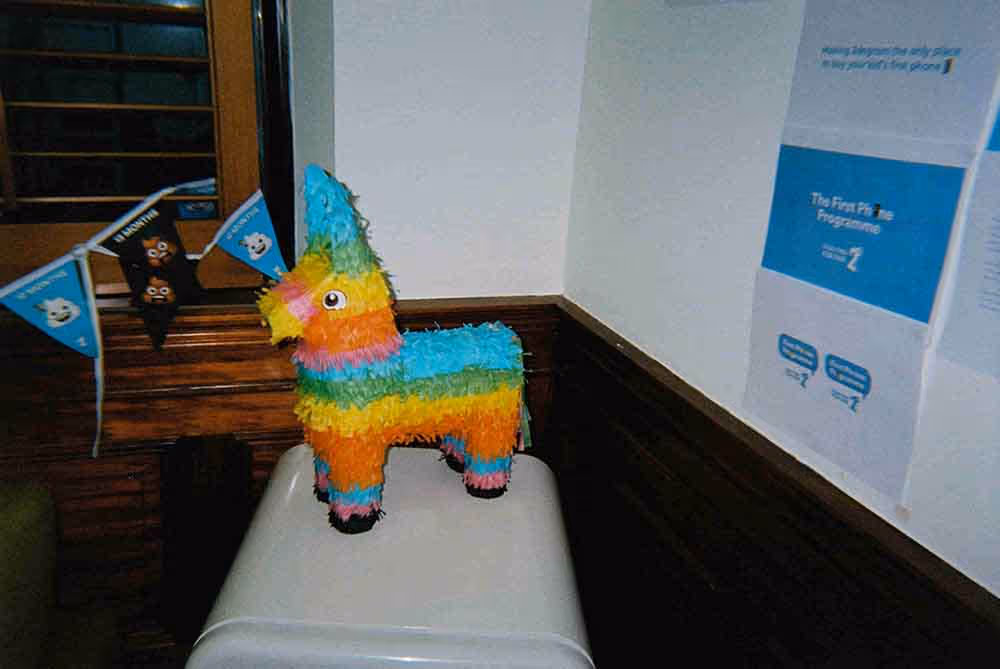 Colourful donkey piñata on a white surface in a room decorated with cartoon-themed bunting and posters for 'The First Pi' Programme.