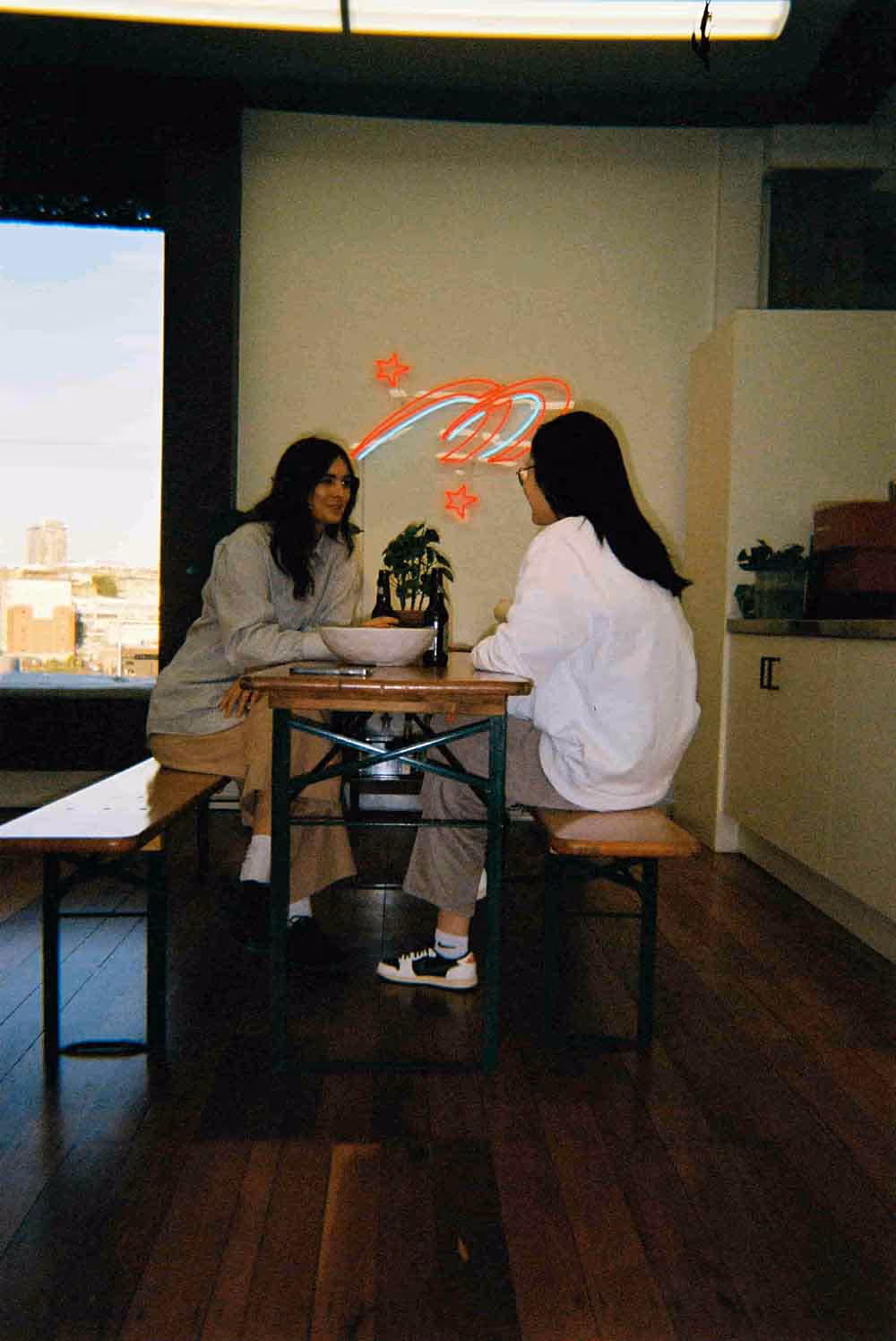 Two people sitting at a wooden bench table in a modern office kitchen area, talking with a plant and bowls on the table, with a red neon star-and-swoosh sign on the wall behind them.