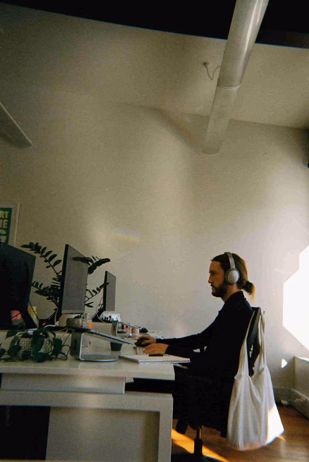 Person with headphones and a tote bag over their chair working at a computer desk in a sunlit office, with indoor plants and minimal decor.