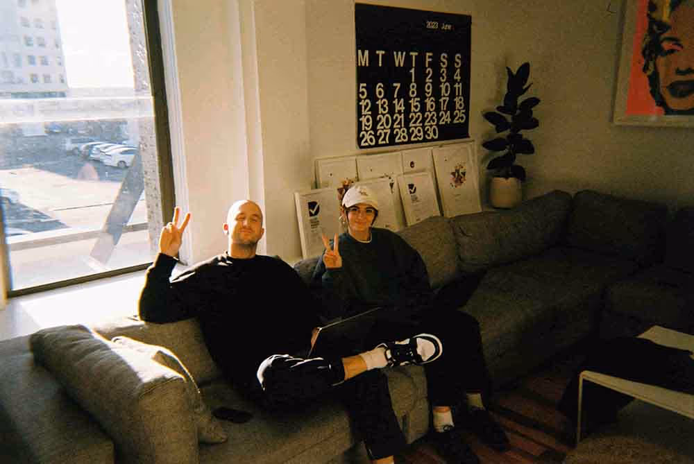 Two people sitting on a grey couch in a sunlit lounge area, both making peace signs toward the camera, with a large wall calendar and framed artwork in the background.