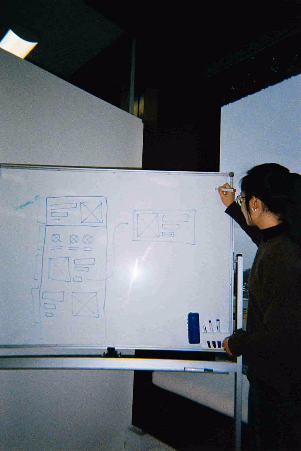 Person sketching a wireframe or website layout on a whiteboard using blue and red markers, with various rectangles representing interface elements and pages.