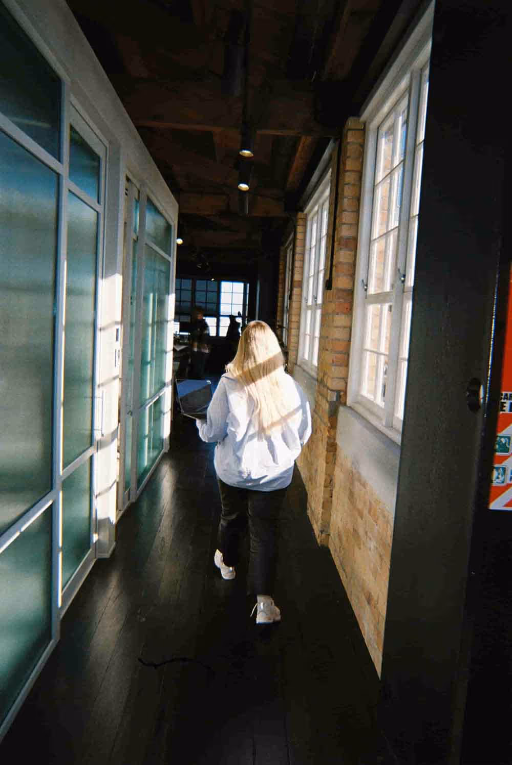 Person walking down a sunlit hallway lined with windows and frosted glass panels, holding a laptop, with warm light casting shadows along the wooden floor and brick walls.