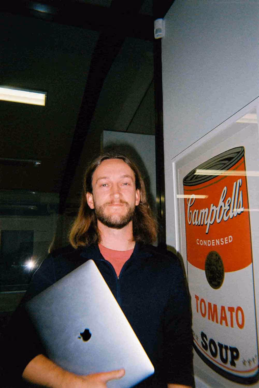 A man with long hair smiles while holding a laptop next to a framed Campbell’s soup poster in a creative office.