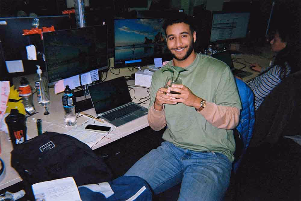 A young man in a creative office setting smiles while holding a plant, representing life inside the advertising industry.