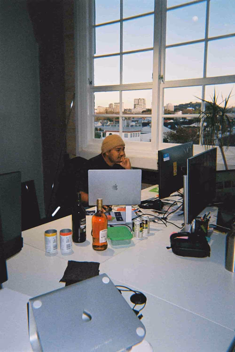Man wearing a beanie focused on work at a desk with multiple monitors and a laptop, large windows behind showing city buildings at sunset, with drinks and cans scattered on the white tabletop.