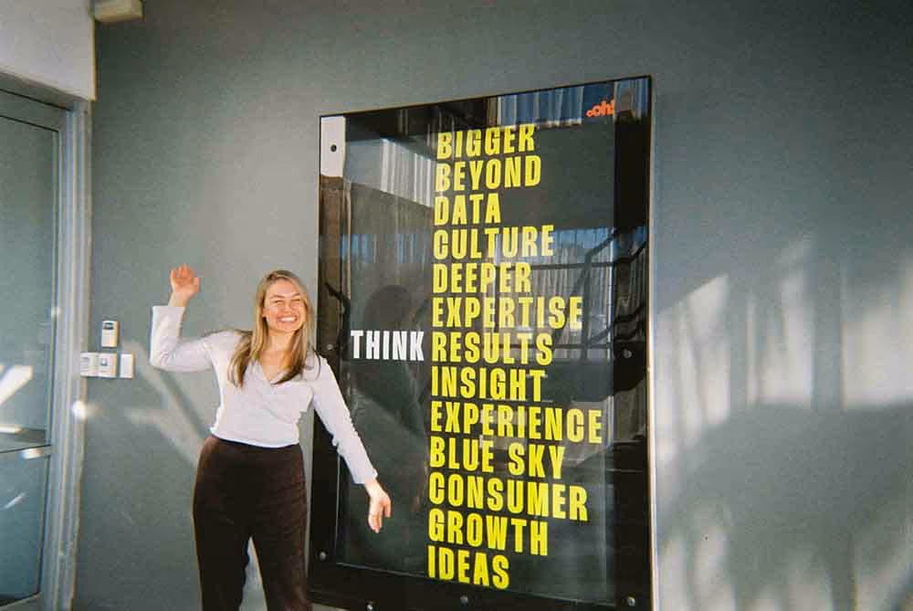 A woman smiling and posing next to a bold agency poster that reads 'Think Bigger, Beyond, Data, Culture…'