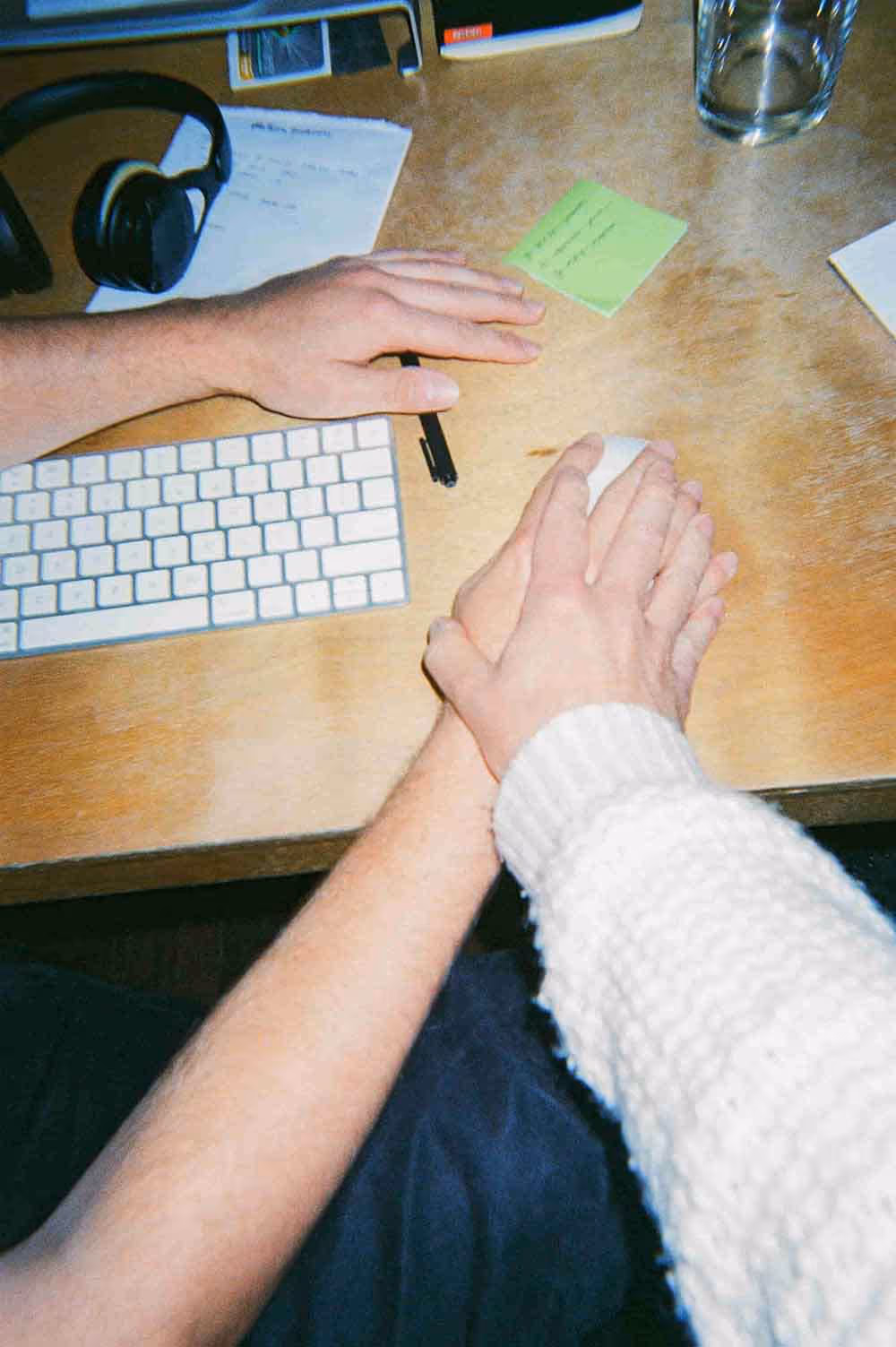 Close-up of two hands overlapping on a computer mouse, next to a keyboard on a wooden desk with scattered notes and a pair of headphones.