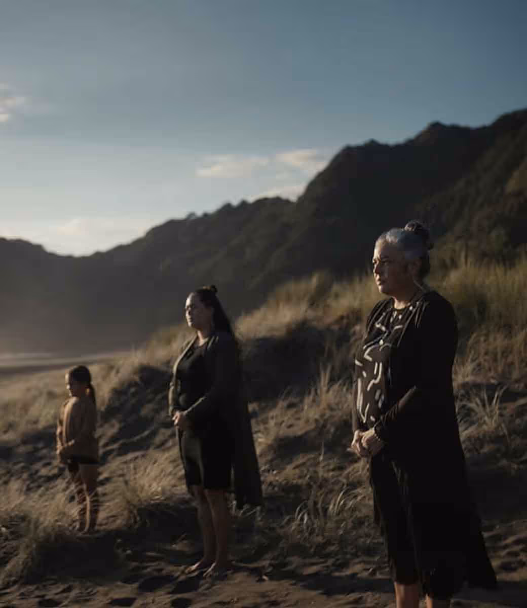 Three generations of Māori women—child, mother, and elder—stand solemnly on a coastal dune at sunset, facing the horizon in a moment of reflection and cultural reverence.