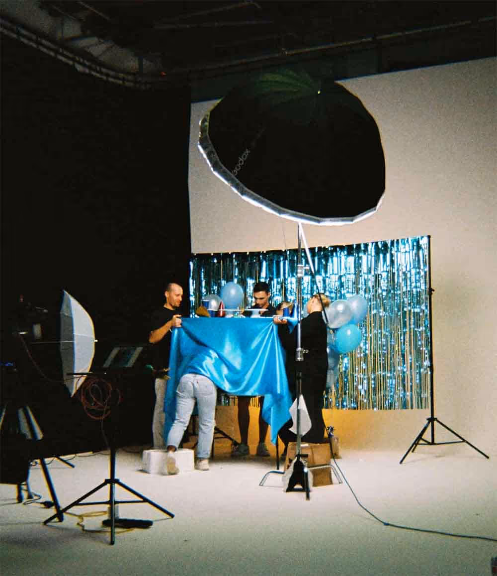 A group of people setting up a photo shoot in a studio, arranging balloons and a shiny blue backdrop under large studio lights.