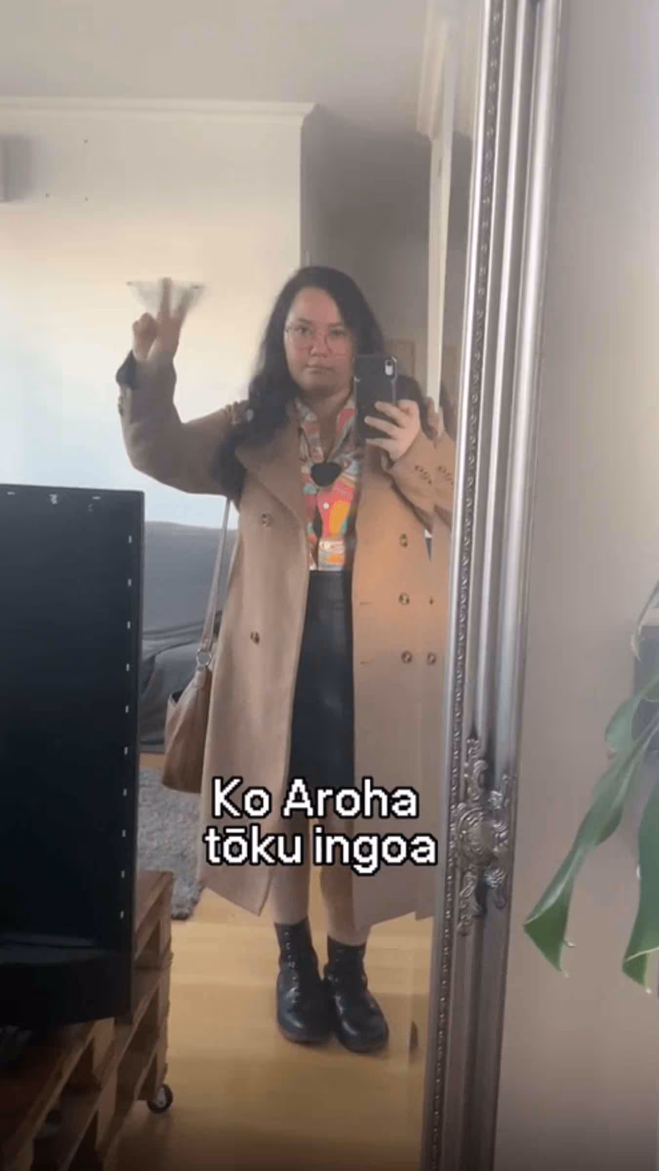 A person taking a mirror selfie in a long coat and boots, flashing a peace sign with the caption 'Ko Aroha tōku ingoa' (My name is Aroha).