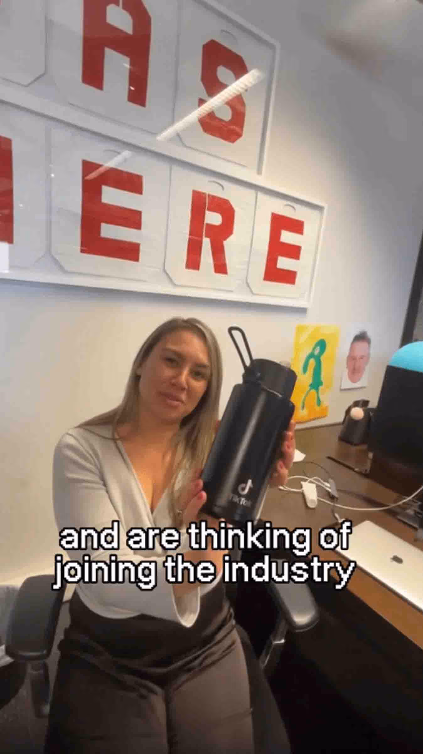 A woman sitting at her desk holding a TikTok-branded drink bottle, with on-screen text that says 'and are thinking of joining the industry'.