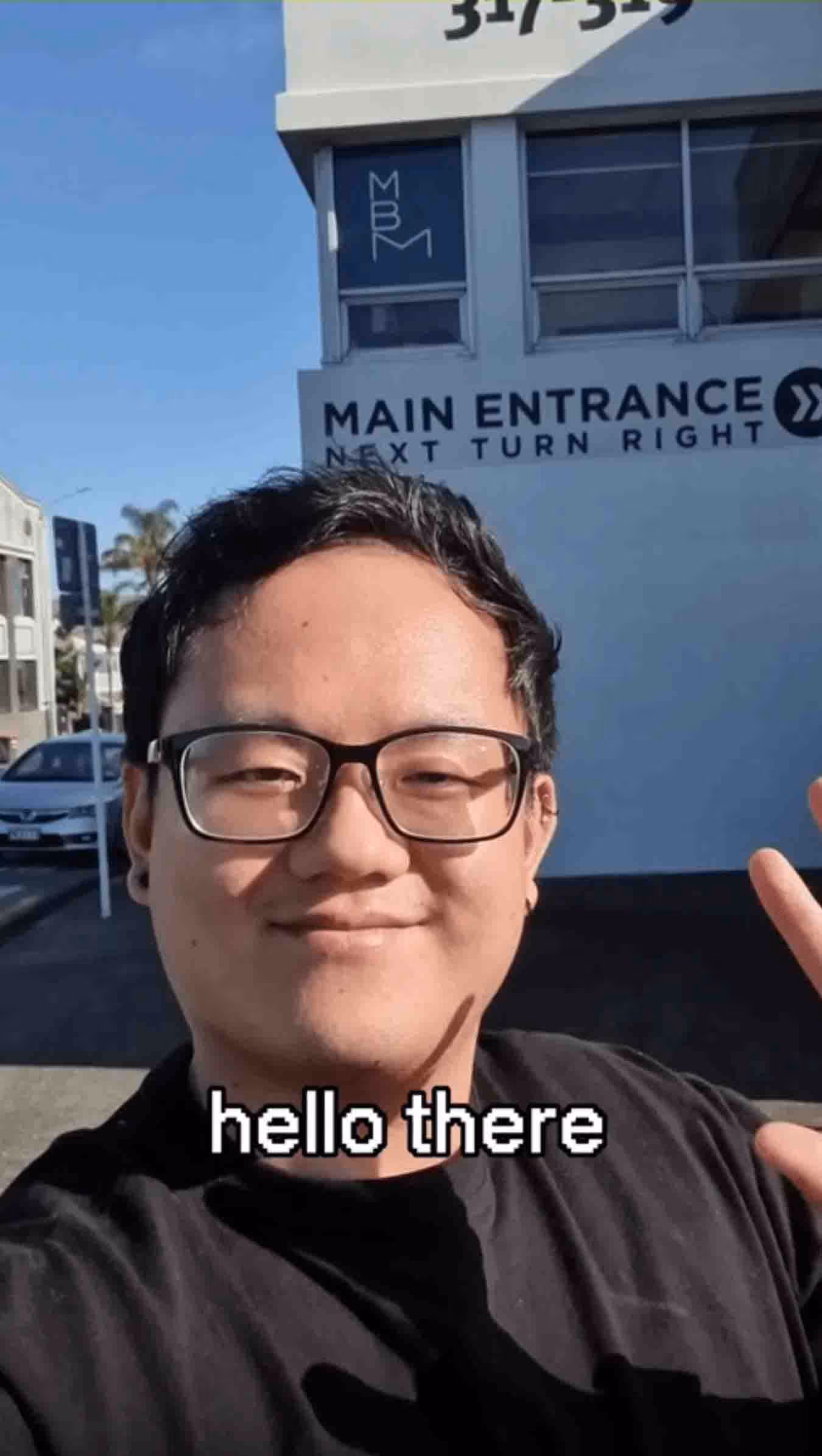 A person smiling and waving outside the MBM office building, with on-screen text that reads 'hello there'.